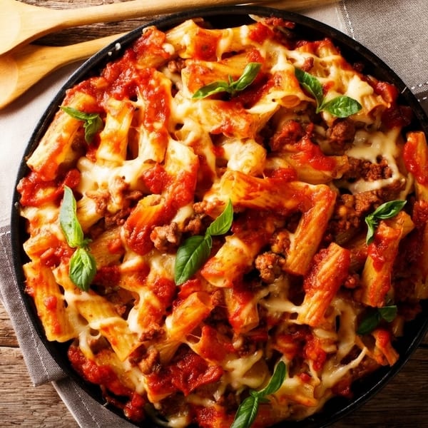 Baked ziti pasta with meat, tomato sauce, melted cheese, and basil in a black pan.